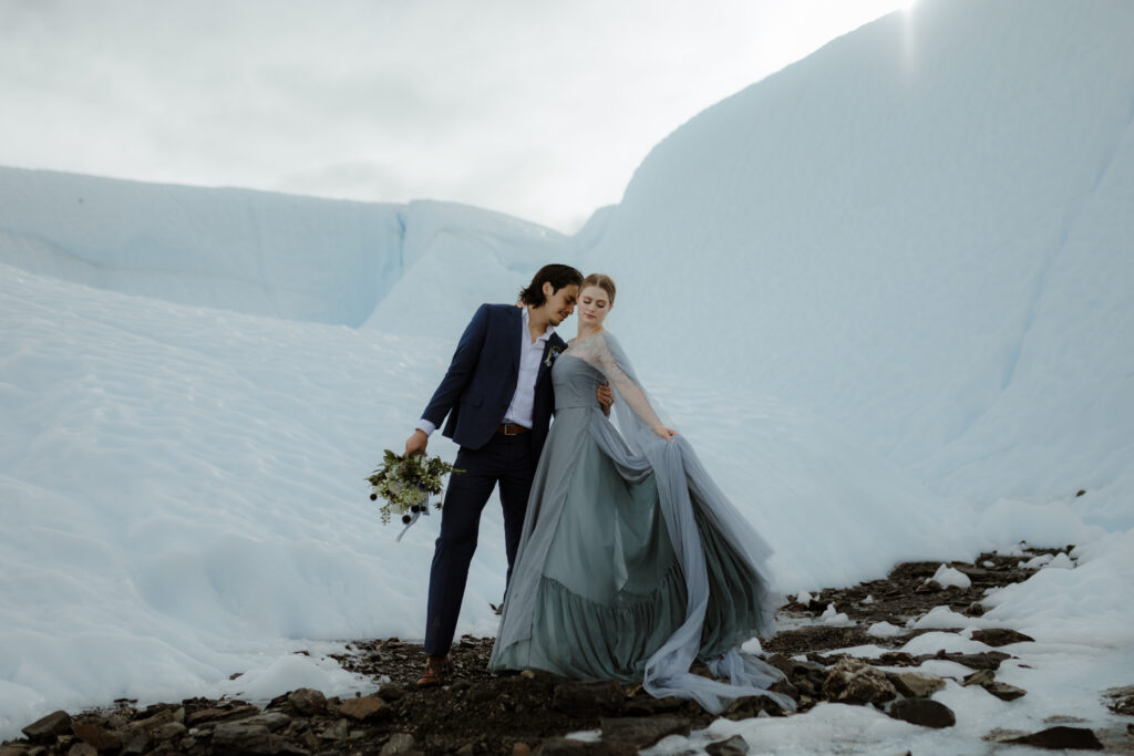 A bride in a blue wedding dress and a groom in a navy suit stand on Matanuska Glacier in Sutton Alaska