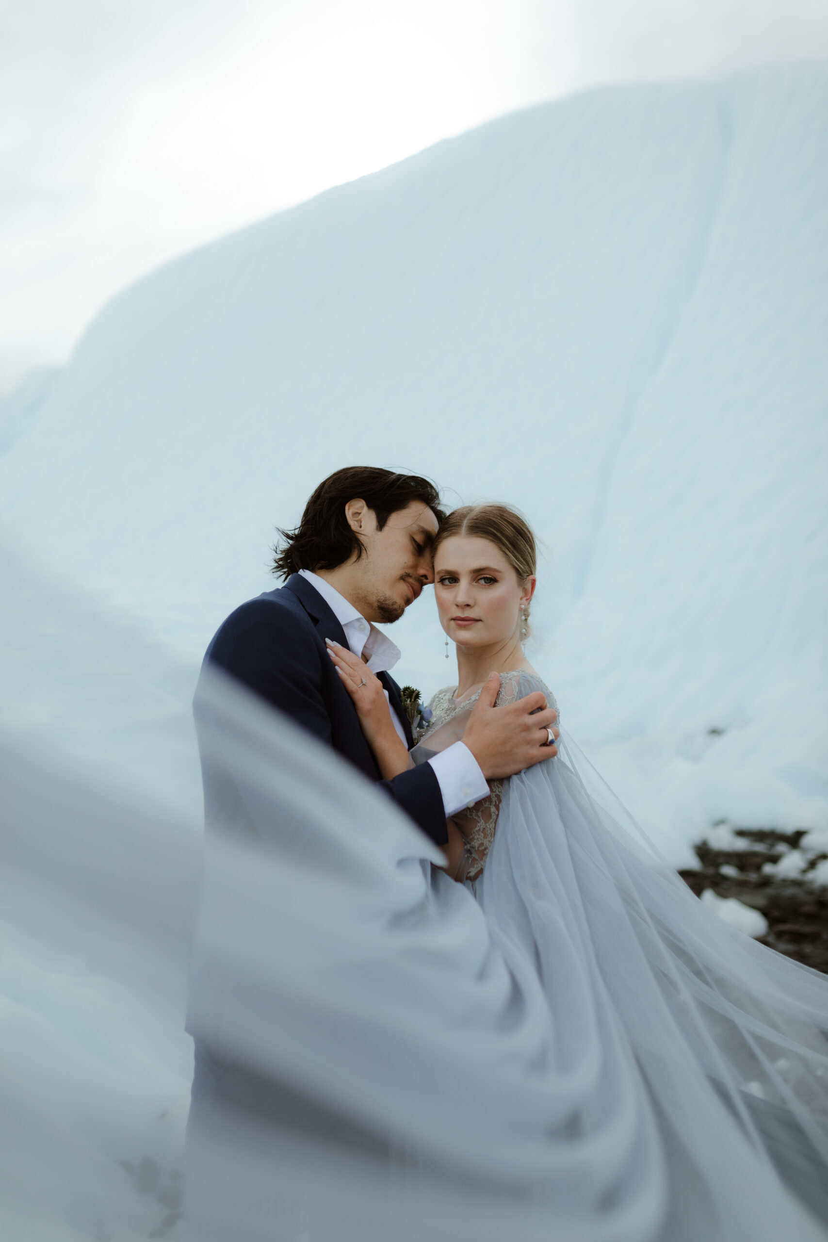 A bride in a blue wedding dress and a groom in a navy suit stand on Matanuska Glacier in Sutton Alaska